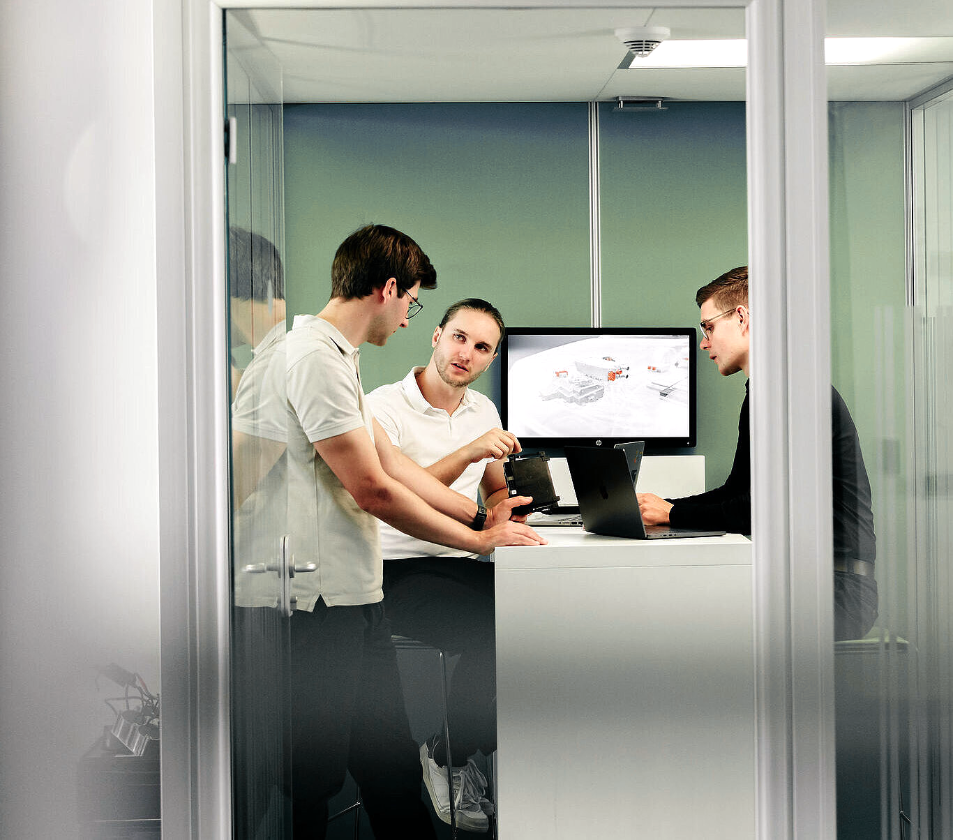 Drei junge Männer besprechen in einem kleinen Glasraum ein technisches Gerät, während auf einem Monitor eine technische Grafik angezeigt wird.