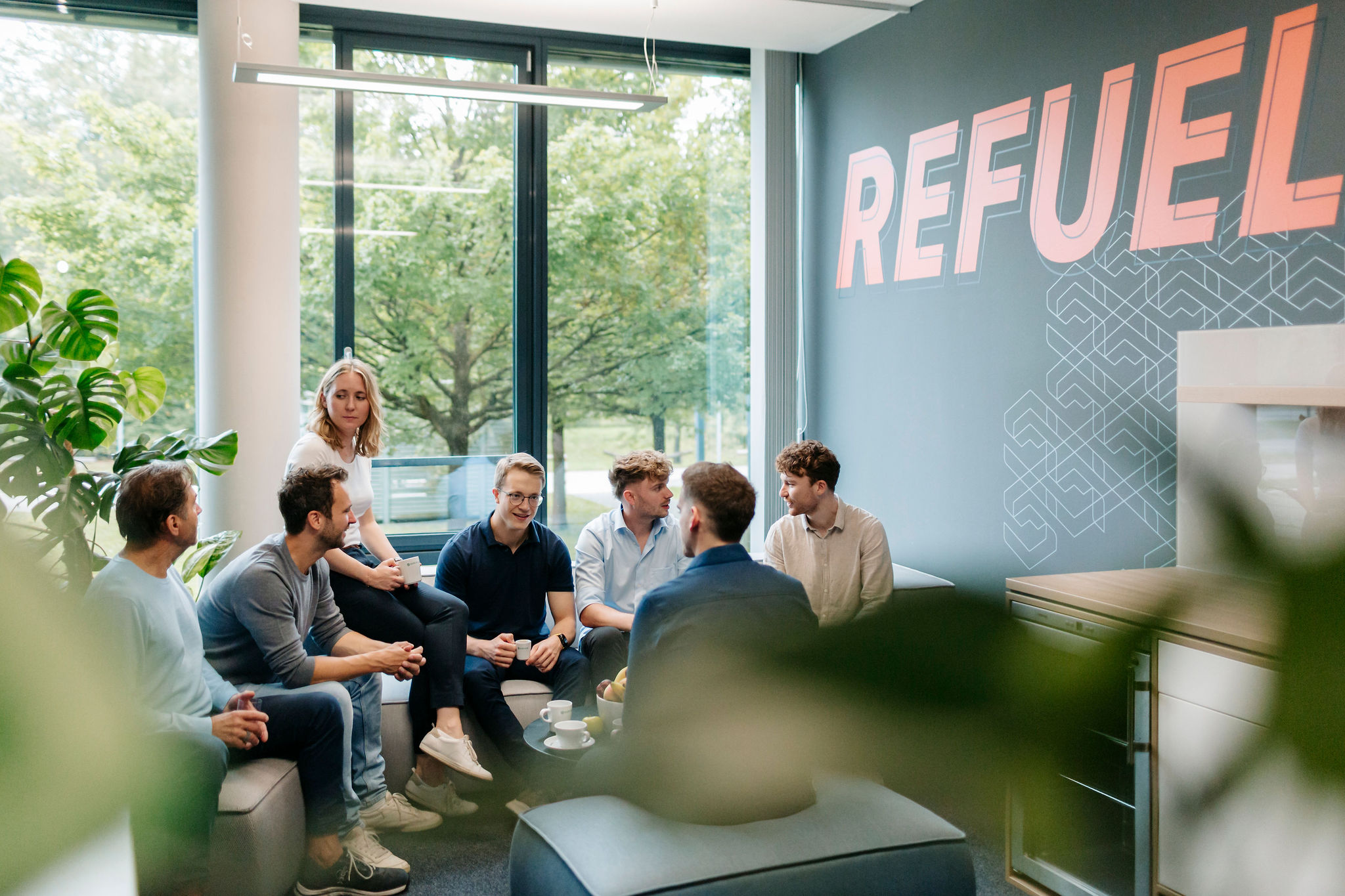 Gruppe junger Fachkräfte sitzt entspannt in einer modernen Lounge mit großer Fensterfront und unterhält sich bei Kaffee.