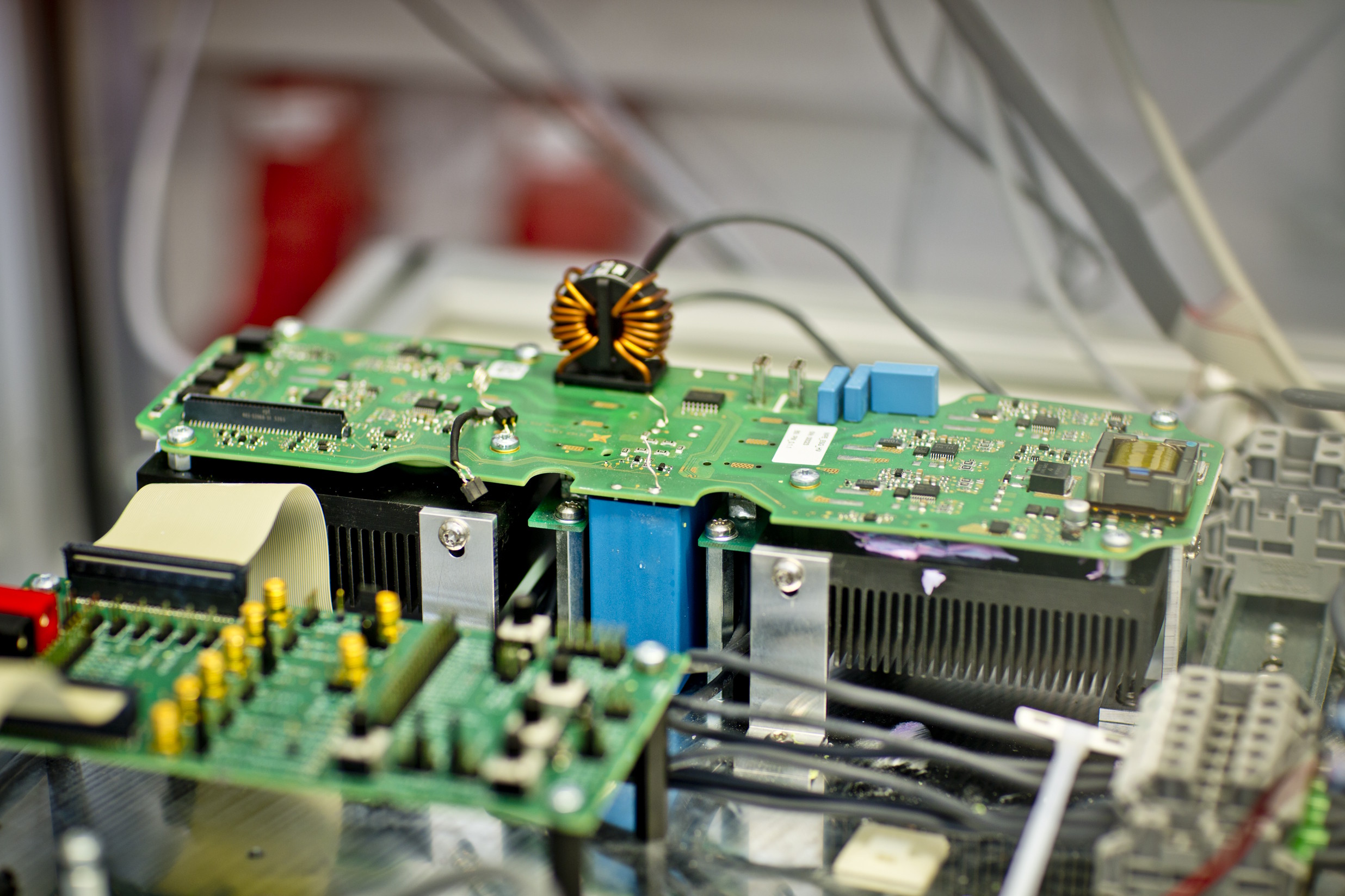 Nahaufnahme einer offenen elektronischen Baugruppe mit Leiterplatten, Bauteilen und angeschlossenen Kabeln auf einem Prüfstand.