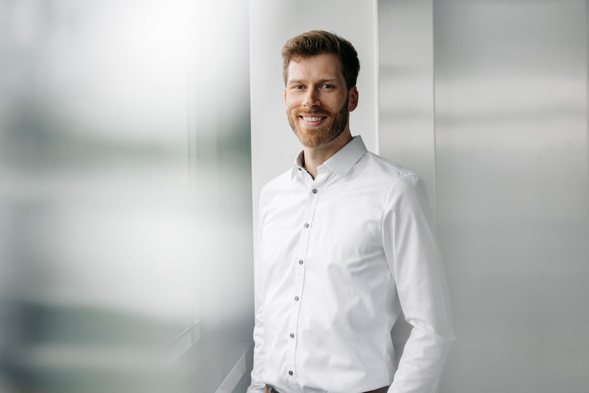 Ein Mann in weißem Hemd steht lächelnd vor einem Fenster in einem hellen Büro.