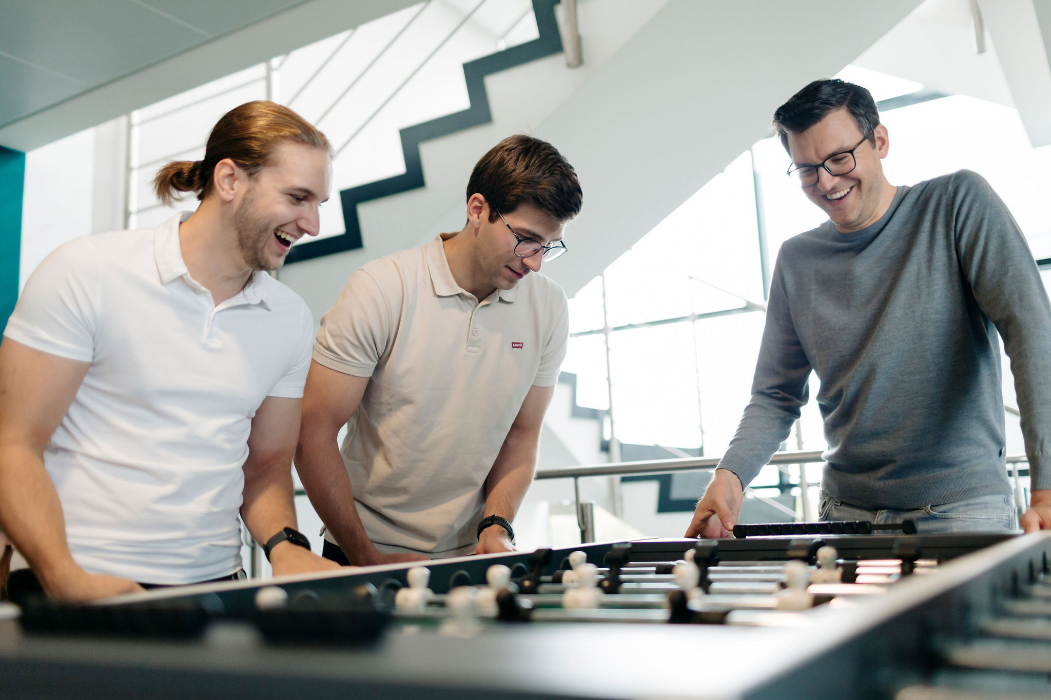 Drei Personen stehen in einem hellen Treppenhaus zusammen an einem Tischkicker und lachen, während sie spielen.